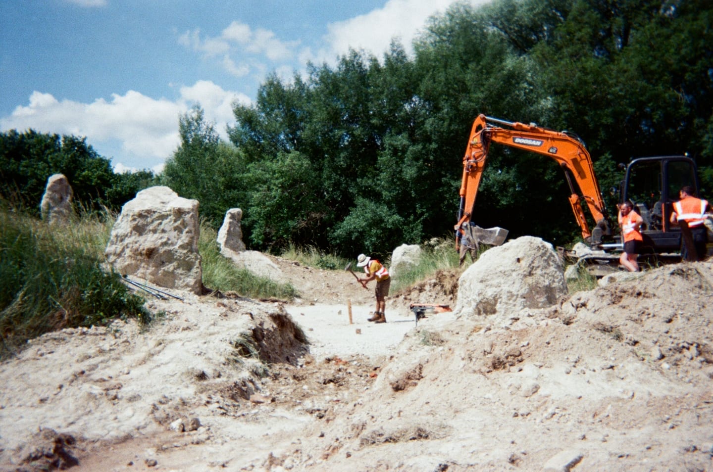 A bright orange digger is digging on a rocky site. There are standing stones around. The rocks are light coloured. There is a person just in front of the digger wearing a sunt hat, hi viz jacket and dark trousers looking at the floor. The Luton Henge standing stones are being installed.