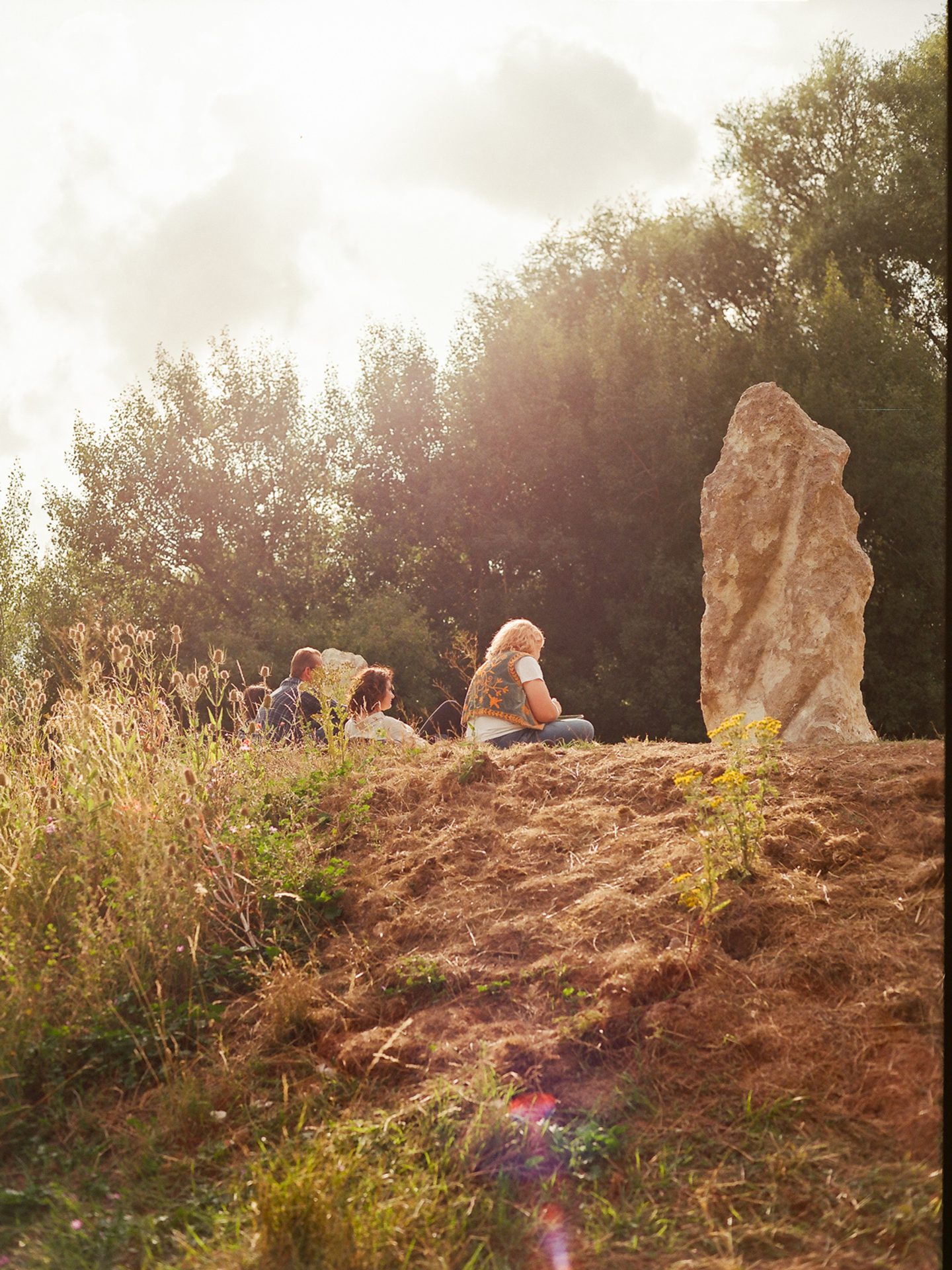 We are looking at a banking with one of the Luton Henge standing stones at the nearside. It is pale brown and has a twisted look to it though it is pointing straight up, further away people are sitting on the banking looking relaxed and facing away from us. The light is amber and it feels like evening, there are trees in the background