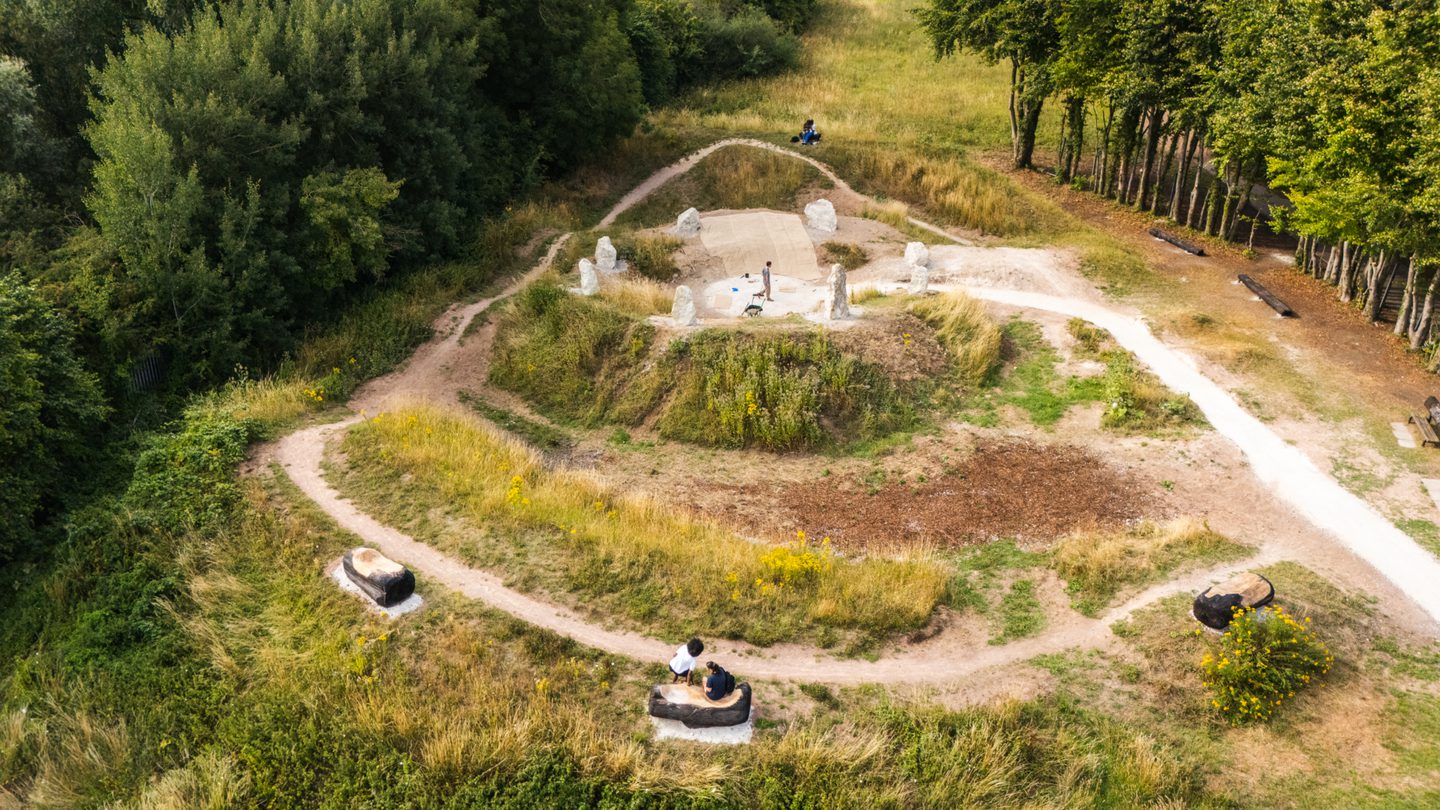 Drone image of Luton Henge with the stones, benches and paths visible. We can see a chalk path leading to standing stones on top of a small mound. Trees surround the mound.