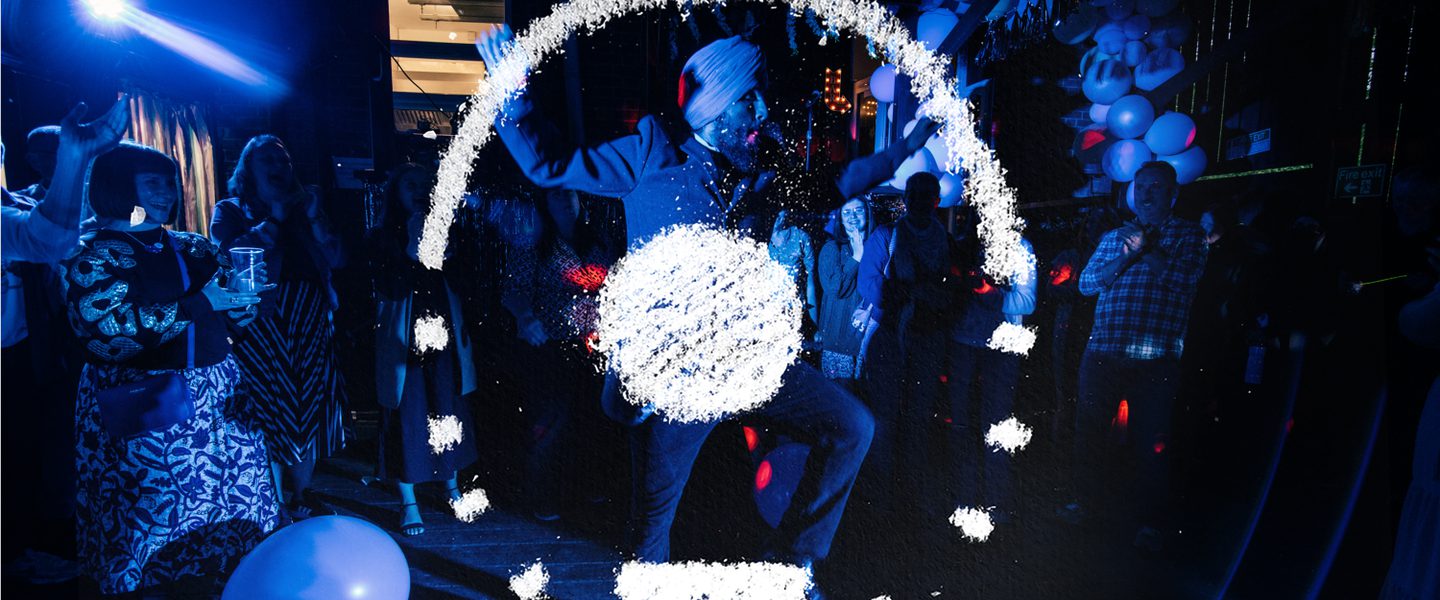People who look to be in a performance, they have hands high. There is a balloon on the floor. The light on them is blue purple. Imposed on the top is a chalk drawing. It's a circle with the top half of the circle a solid line and the bottom dotted. At the bottom of the circle is a sold rectangle making up part of the line of the circle. In the middle is a solid circle.
