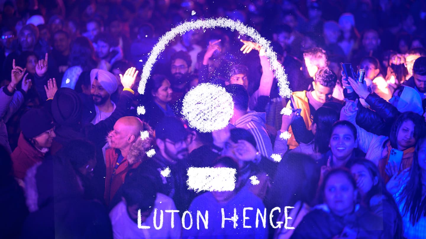 A packed room full of people dancing, we can see heads and shoulders of people standing close to each other. The light on them is blue purple. Some hands are lifted. Imposed on the top is a chalk drawing. It's a circle with the top half of the circle a solid line and the bottom dotted. At the bottom of the circle is a sold rectangle making up part of the line of the circle. In the middle is a solid circle. Underneath, written in chalk 'Luton Henge'.