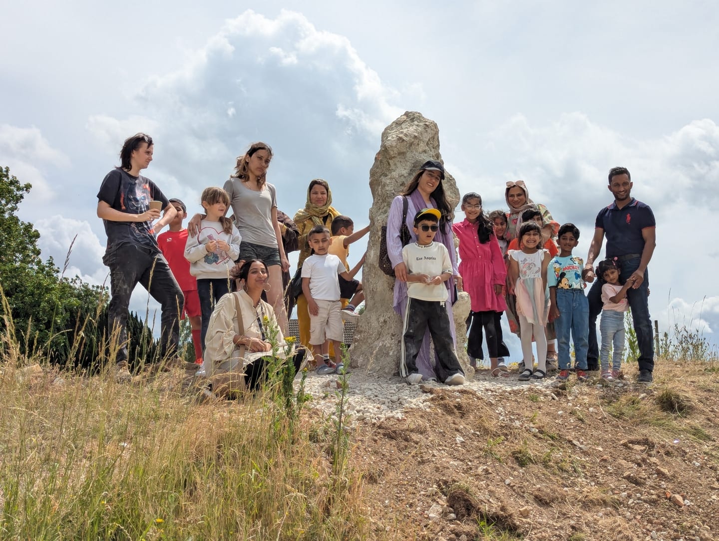 A group of people of all ages stand next to a standing stone. The stone is chalk, a pale rock, it has a point at the top and is on top of a banking. The people all look happy and celebratory.
