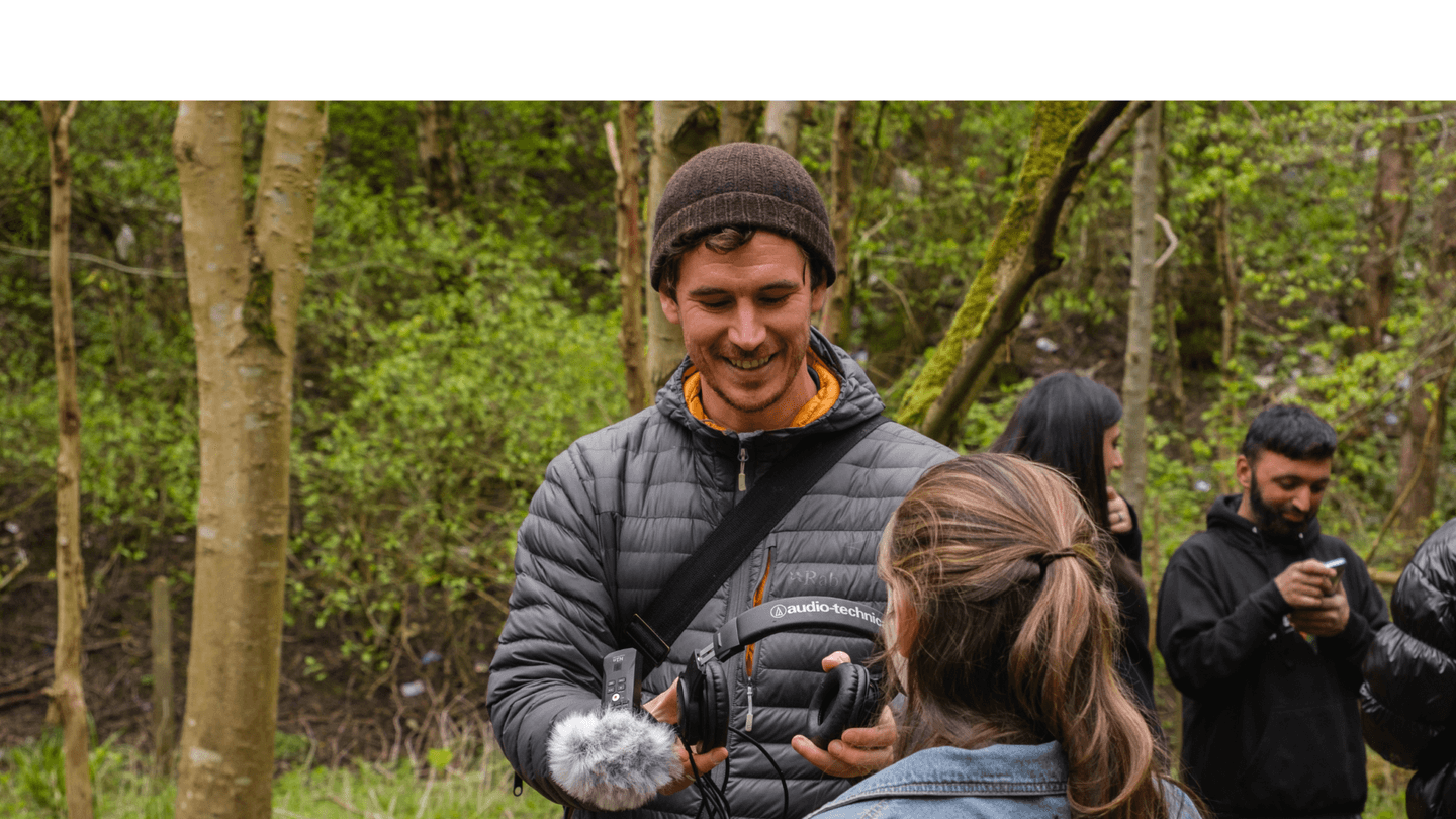 A man in a padded jacket is handing a pair of headphones to a girl with long hair and a denim jacket. They are outside in the woods. Behind them other people are talking.