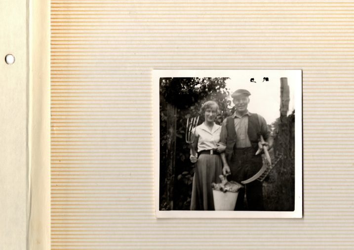 Sam and Beryl
A small square photograph with a white trim, stuck to the page of a photo album. The page is covered in horizontal orange lines, faded and weathered with age. On the left hand side is a cream strip with a hole punch, designed to fit into an album. The photograph itself is black and white, and shows a young woman holding the arm of an older man. Behind them sits foliage, perhaps part of a farm.
The young woman, Beryl, holds a pitchfork in one hand, and the arm of her father with the other. She wears a long circle skirt, belted with a large clasp, and a white shirt. Her hair is short and slightly curled. Sam wears a shirt and an open waistcoat. He holds a bucket of what may be cabbages in one hand, and a scythe and rake in the other. His trousers are high waisted, his shirt is tucked in, and atop his head sits a flat cap. Both are smiling.
I chose to include this photograph because Beryl, my Grandmother and Sam’s daughter, gave me the inspiration for the title of the poem ‘listen to them cows beeling’. She told me of the phrases Sam used to say often, and this one stuck with me.