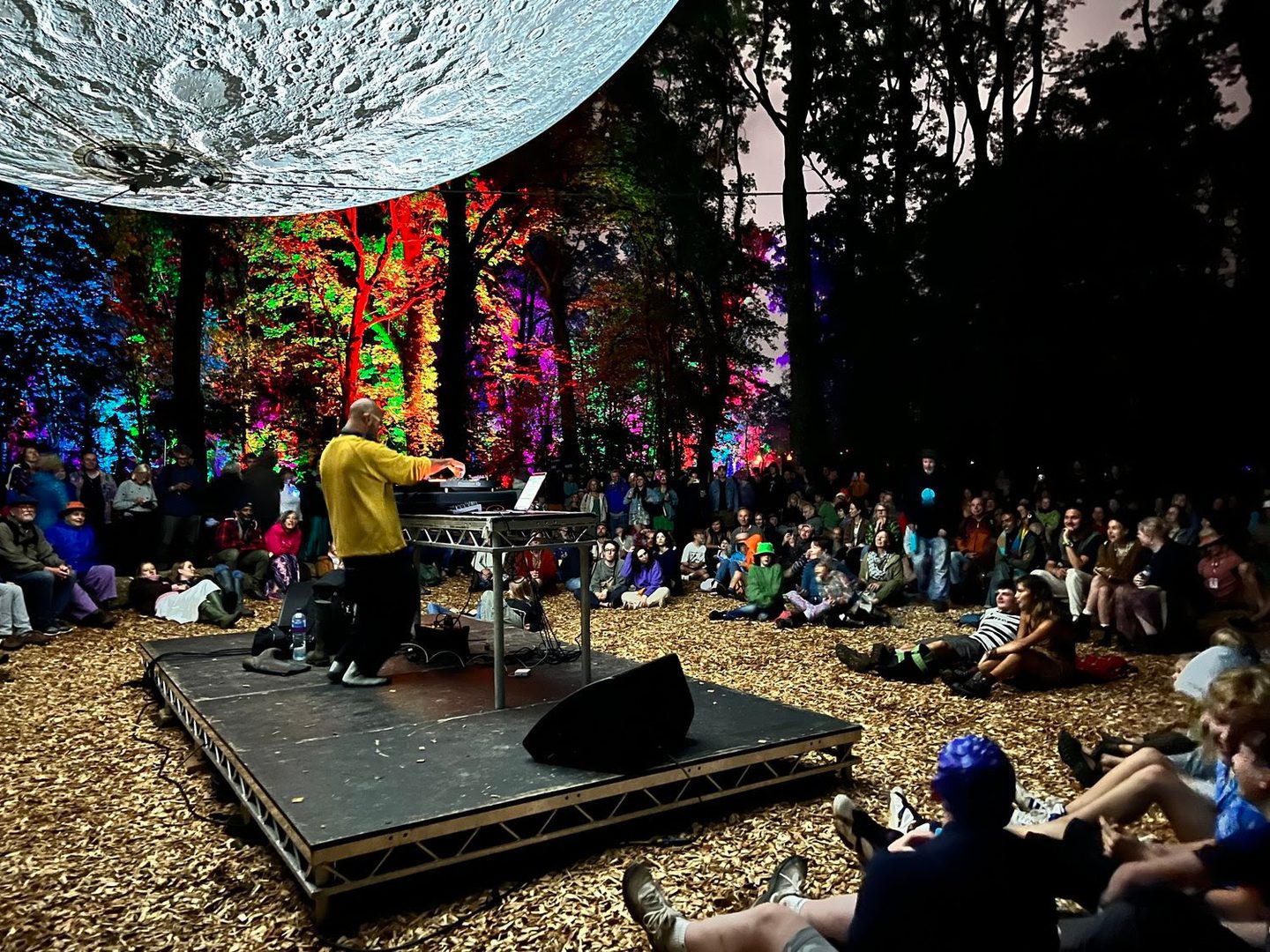 A man DJs on a stage in the woods. Above him is a huge model of the moon. People sit listening. The trees behind are lit with brightly coloured lights.