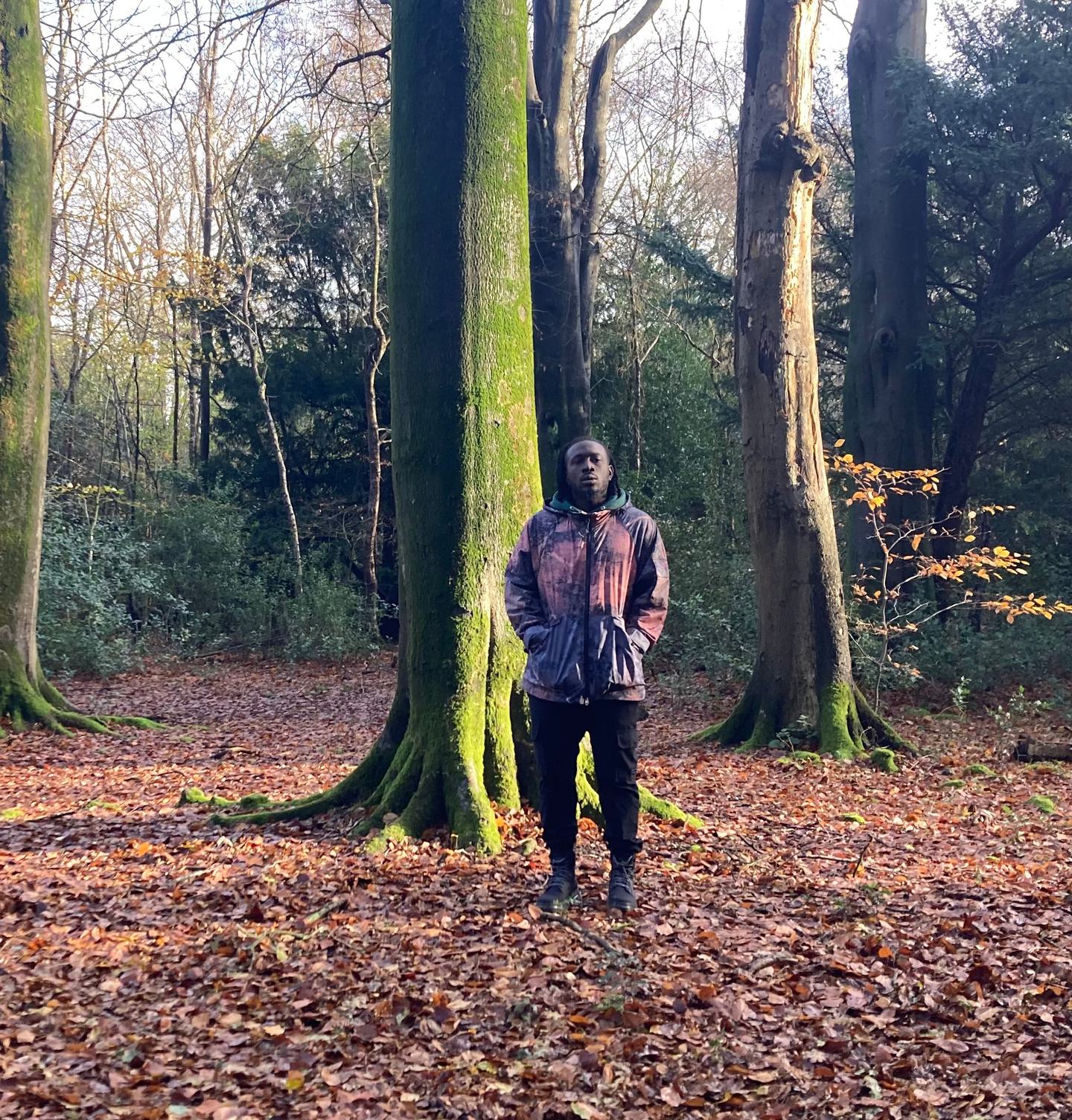 Still Shadey is standing in front of a tree looking directly at the camera. He has locks and is wearing a jacket and a pair of black jeans. He looks thoughtful. The tree is tall and straight and has moss on its trunk. The floor is covered in autumn leaves.