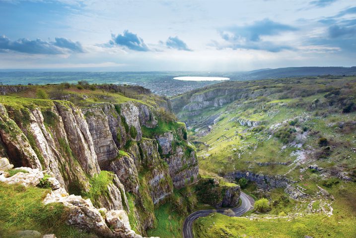 A view of Cheddar Gorge, vertical cliffs with a long view out to a sparkling lake in the far distance.