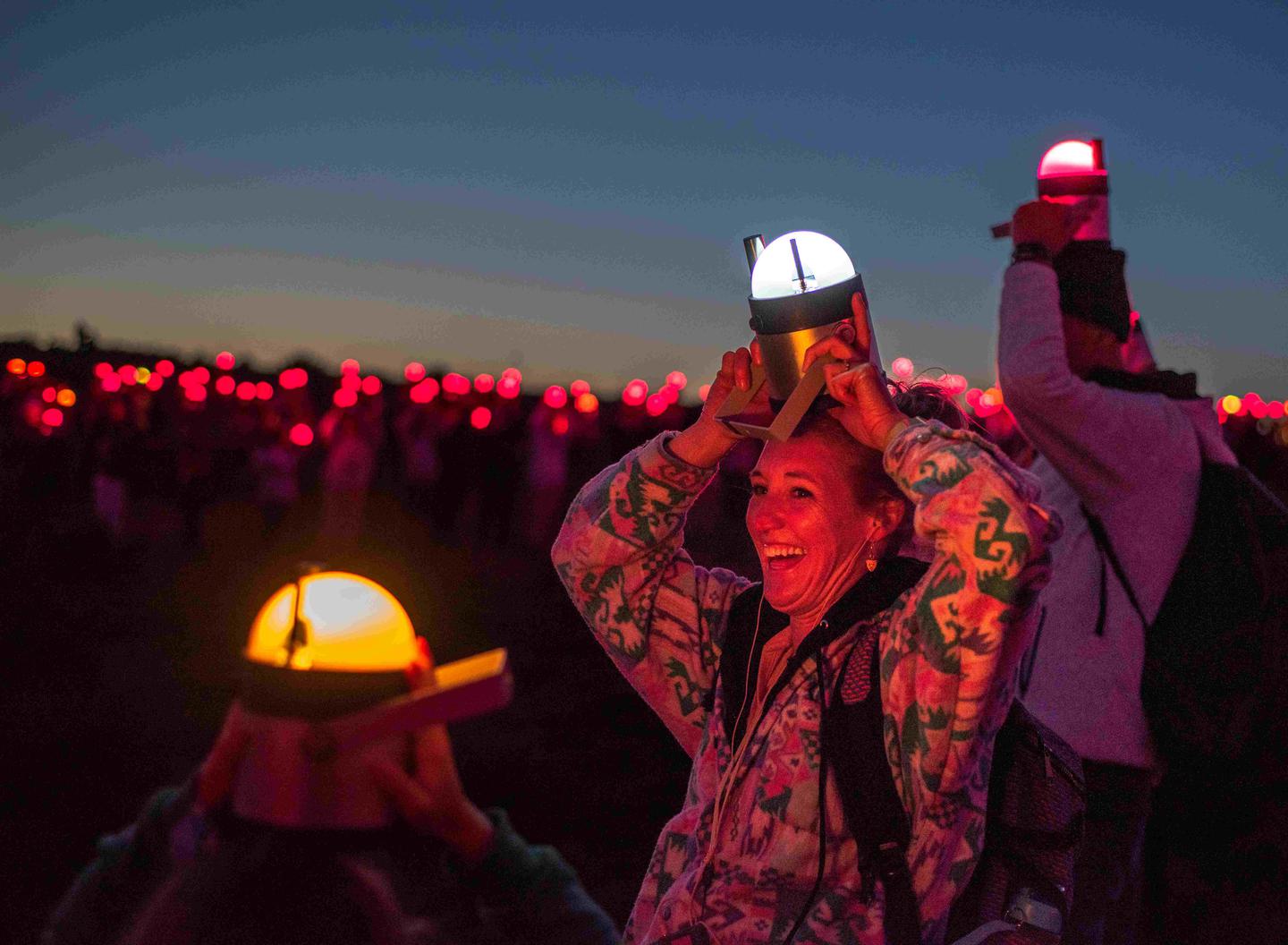 People stand with lanterns above their heads. In the foreground we see a family with yellow and white lanterns. Behind them are hundreds of people with red lanterns.