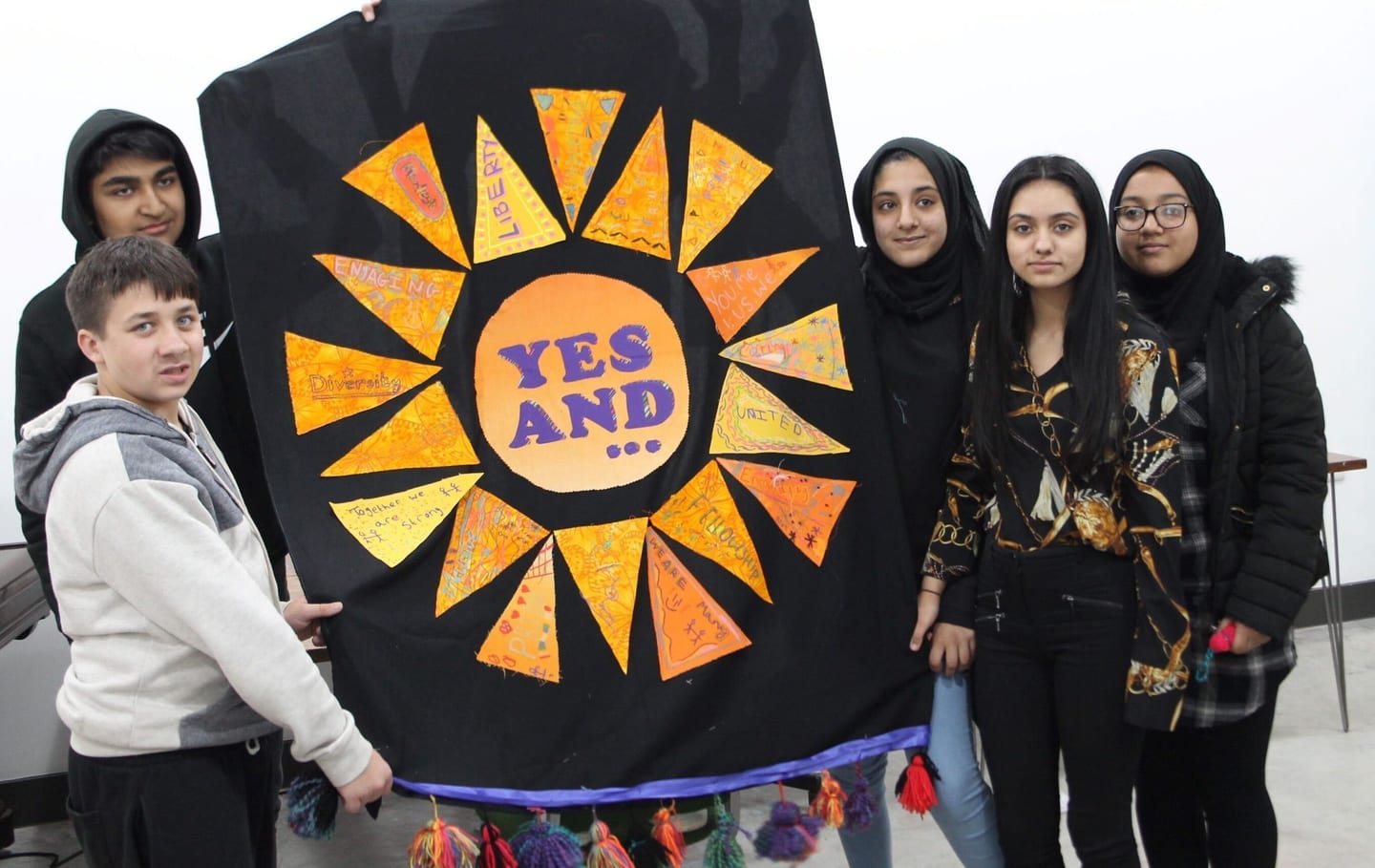 Six young people stand with a beautiful homemade banner. It has a black background and a yellow geometric sunshine shape that says 'Yes And' on it.