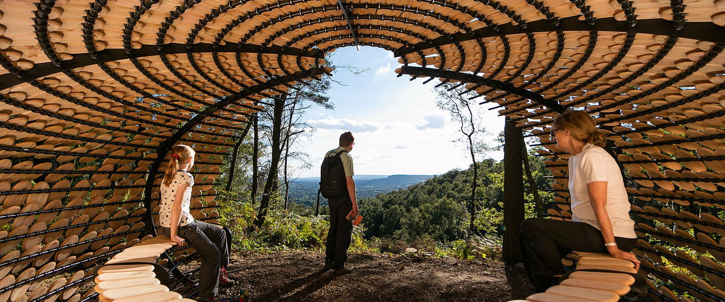 Two people sit on a semi circular bench in a shelter, it is all made of wood and very beautiful. They look out on to a view through trees across a verdant valley.